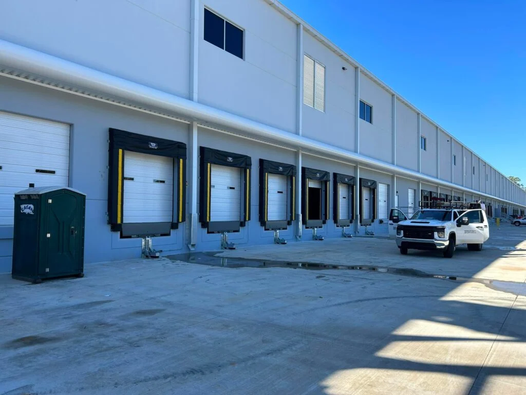 Row of commercial loading dock doors with dock seals and levelers outside a large warehouse building, with a work truck parked nearby.
